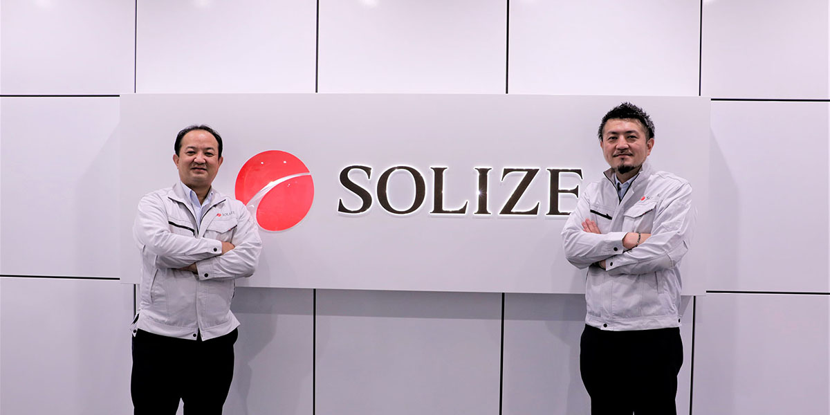Yoshihiro Nomura and Tohru Ota stand in front of the SOLIZE logo
