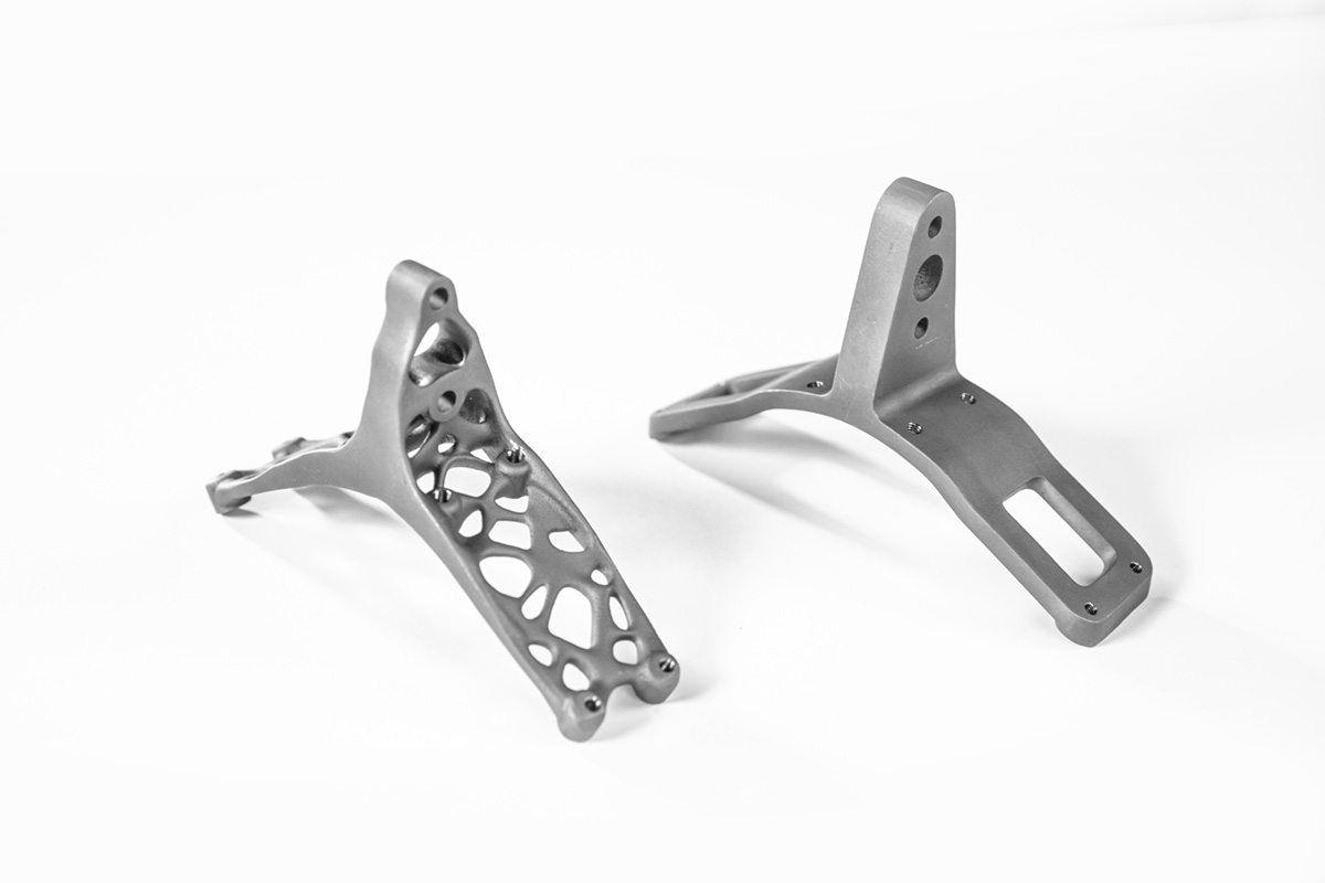 Two titanium brackets side by side. A lightweight version on the left is based on generative design and has irregular holes